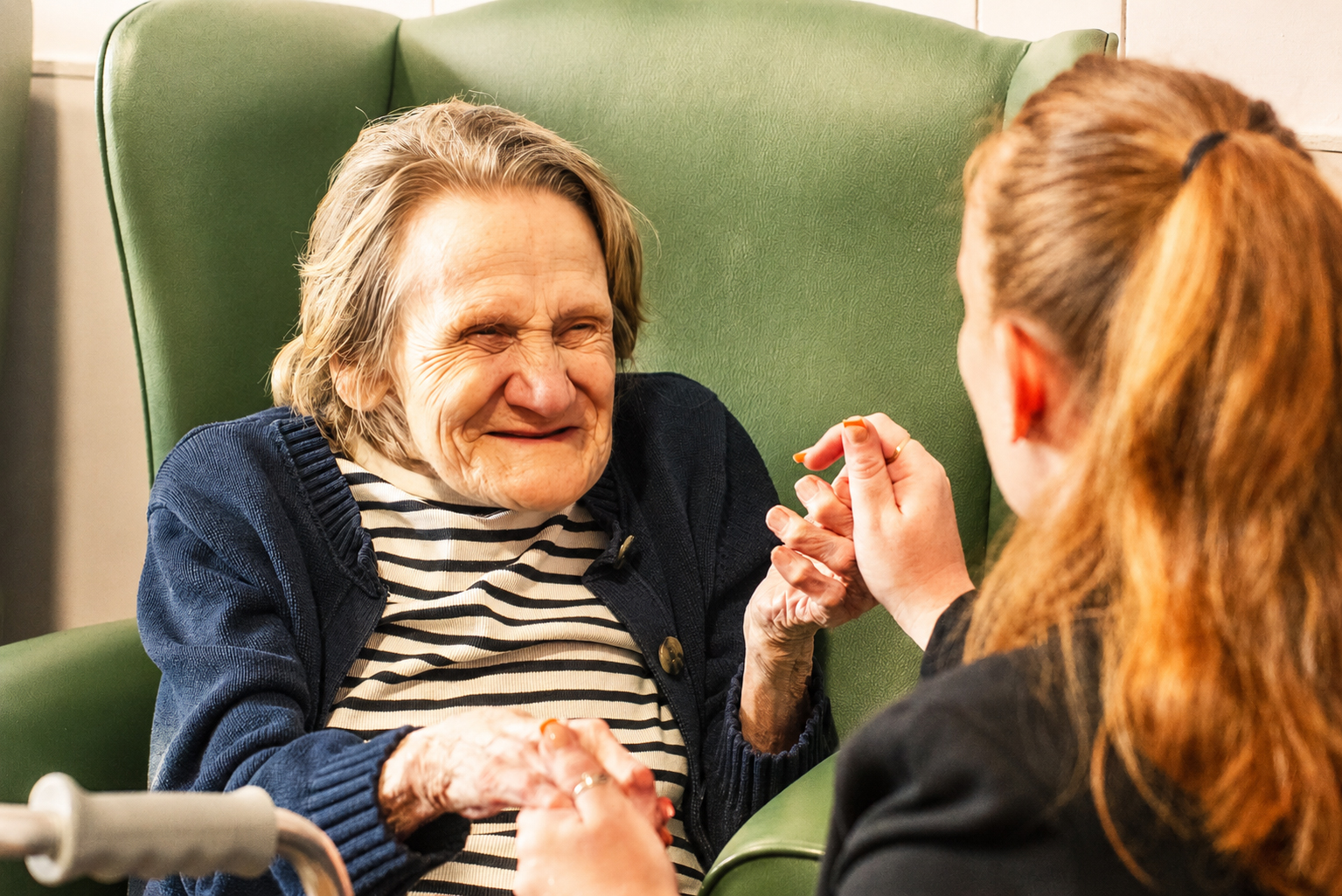 An elderly woman in a striped shirt and blue cardigan sits in a green armchair, smiling and holding hands with a younger woman with reddish-brown hair, who faces her and smiles warmly.