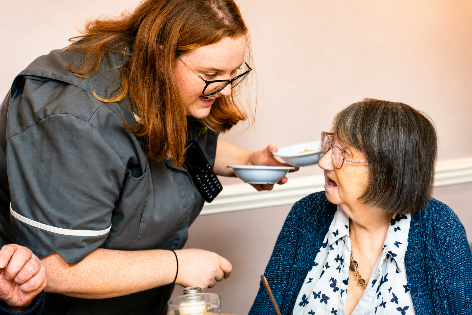 A carer in uniform smiles and serves food to an elderly woman with glasses, who is sitting at a table and looking up at her, both appearing engaged in conversation.