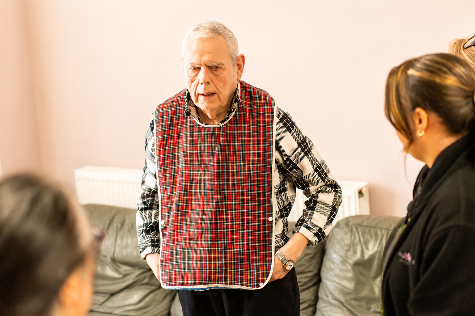 An older man wearing a checked adult bib stands in a lounge at Park Lane Care Home, facing three people who are partially visible in the foreground. He appears to be engaged in conversation with them.